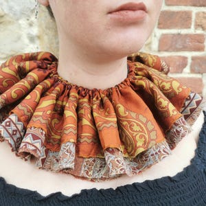 Könnte beinhalten: Ein dekorativer Rüschenkragen aus Stoff in Orange-, Gelb- und Brauntönen mit einem Muster im Paisley-Stil. Der Kragen ist am Halsausschnitt gerafft und hat einen dekorativen Rand. Das Kleidungsstück wird über einem schwarzen Oberteil getragen, mit einer Backsteinmauer im Hintergrund.