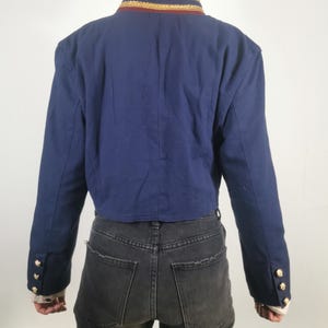 May include: A cropped navy blue blazer with gold trim around the collar and gold buttons on the cuffs. The blazer is worn over a pair of black jeans.