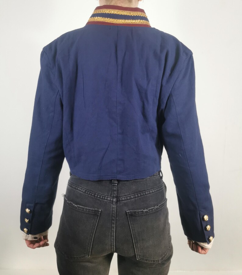 May include: A cropped navy blue blazer with gold trim around the collar and gold buttons on the cuffs. The blazer is worn over a pair of black jeans.