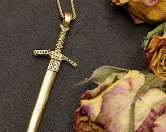 Pode incluir: Um colar com pingente dourado com um design de espada. O pingente de espada tem detalhes intrincados na empunhadura e na lâmina. O colar é exibido em uma superfície escura com rosas secas ao fundo.