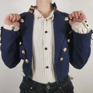 May include: A navy blue cropped jacket with gold buttons and a white shirt underneath. The jacket has a collar with a gold and red stripe.