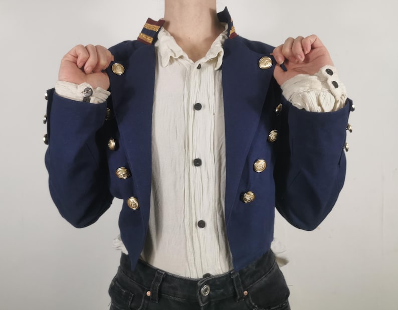 May include: A navy blue cropped jacket with gold buttons and a white shirt underneath. The jacket has a collar with a gold and red stripe.