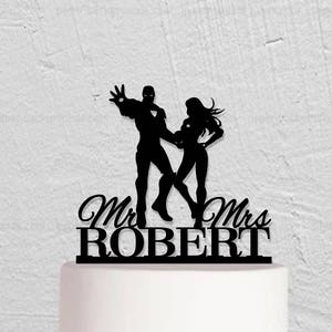 May include: A wedding cake topped with a black silhouette of a couple, with the words "Mr & Mrs ROBERT" underneath. The cake is decorated with a floral arrangement of pink, white, and purple flowers.