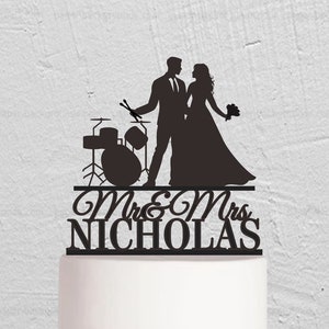 May include: Black silhouette cake topper of a bride and groom with a drum set. The groom is holding drumsticks and the bride is holding a bouquet. The text "Mr. & Mrs. Nicholas" is below the couple.