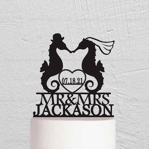 Op de afbeelding: Zwarte silhouet van twee zeepaardjes die elkaar kussen met een hartje waarop "07.18.21" staat en de tekst "MR & MRS JACKSON" eronder. De topper staat op een witte en gouden bruidstaart die versierd is met roze en paarse bloemen.