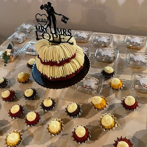 May include: A two-tiered red velvet cake with white frosting and a black silhouette of a couple with the text "Mr. & Mrs. Lopez" on top. The cake is surrounded by small cupcakes with white and yellow frosting.