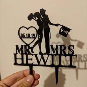 May include: Black silhouette of a couple kissing with a heart and the date 05.10.19. The text "MR. & MRS. HEWITT" is below the couple. The couple is holding a flag that says "V+N".