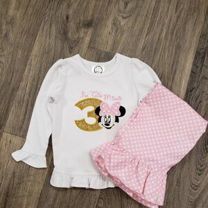 May include: White long-sleeved shirt with a gold glitter number 3 and a pink Minnie Mouse appliqué. The shirt has a ruffle trim. Pink polka dot trousers with a ruffle trim.