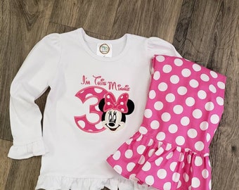 May include: White long-sleeved shirt with a pink Minnie Mouse design and the text "I'm This Minnie 3". The shirt has a ruffled hem. Pink and white polka dot leggings with a ruffled hem.