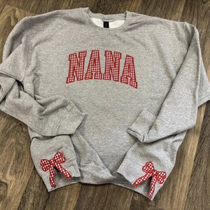 May include: A gray sweatshirt with the word "NANA" in red gingham letters across the chest. The sleeves have red gingham bows at the cuffs. The sweatshirt is made of a soft, comfortable material.