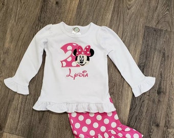 May include: A white long-sleeved shirt with a pink and black Minnie Mouse design and the number 2. The shirt has a ruffled hem and is paired with pink polka dot pants with a ruffled hem. The shirt is personalized with the name "Lydia".