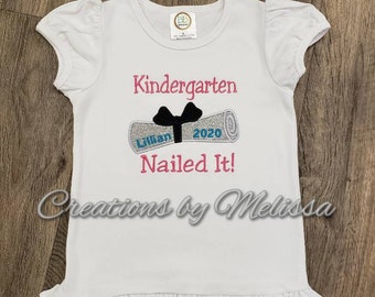 May include: White t-shirt with a pink and black embroidered design that says "Kindergarten Nailed It!" with a graduation cap and the year 2020. The shirt has a ruffled hem and short sleeves.
