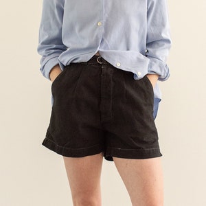 May include: A light blue button-down shirt and black shorts. The shorts have a belt and are paired with black espadrilles. The outfit is casual and suitable for warm weather.