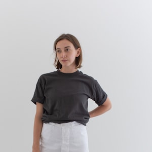 以下が含まれることがあります： 濃いグレーのTシャツと白いワイドパンツを着用した人。Tシャツは半袖でクルーネックです。パンツはハイウエストでボタン留めです。その人は黒い靴を履いています。