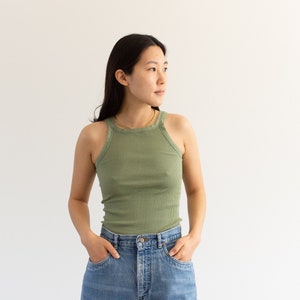 Könnte beinhalten: Eine Frau in einem grünen Tanktop und blauen Jeans, die vor einer weißen Wand steht.