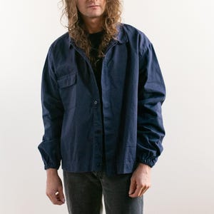 Può includere: Una camicia blu navy con bottoni, due tasche sul petto e maniche lunghe. Le maniche sono raccolte ai polsi. La camicia è indossata sopra una maglietta nera. La persona indossa jeans grigi.