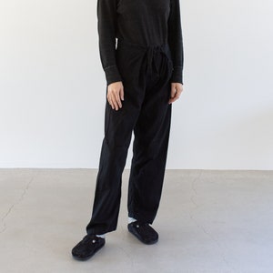 May include: A woman wearing a black long-sleeved shirt and black pants with a tie at the waist. She is standing on a gray floor in front of a white wall.
