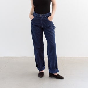 May include: A woman wearing a black tank top and dark blue denim carpenter pants. She is standing with her hands in her pockets and looking to the side.