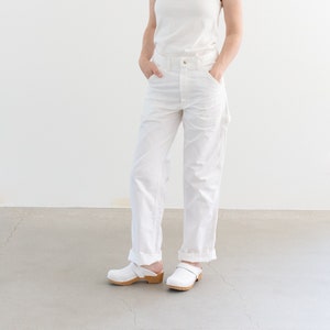 May include: A woman wearing a white tank top and white carpenter pants with a cuffed hem. She is wearing white leather clogs with wooden soles.