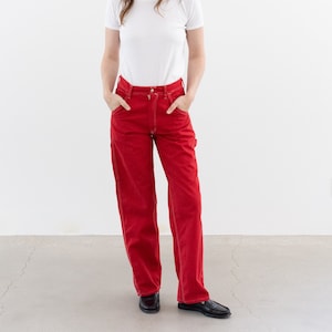 May include: A woman wearing a white t-shirt and red carpenter jeans. She is standing in front of a white wall.