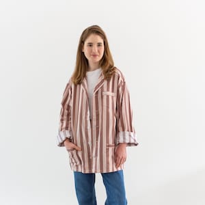 May include: A button-down, striped shirt in shades of red, brown, and cream, paired with blue jeans. The shirt has a relaxed fit and is worn over a white t-shirt. The jeans are cuffed at the ankles, and the person is wearing gray boots.