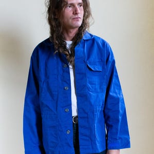 May include: A vibrant blue long-sleeve jacket featuring a classic collar, a front button closure, and a single chest pocket on the left side. The jacket is constructed with a structured fit, long sleeves, and visible stitching details along the front placket.