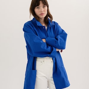 May include: A woman wearing a long, royal blue work jacket with a white button-down shirt underneath. She is wearing white jeans and has her arms crossed.