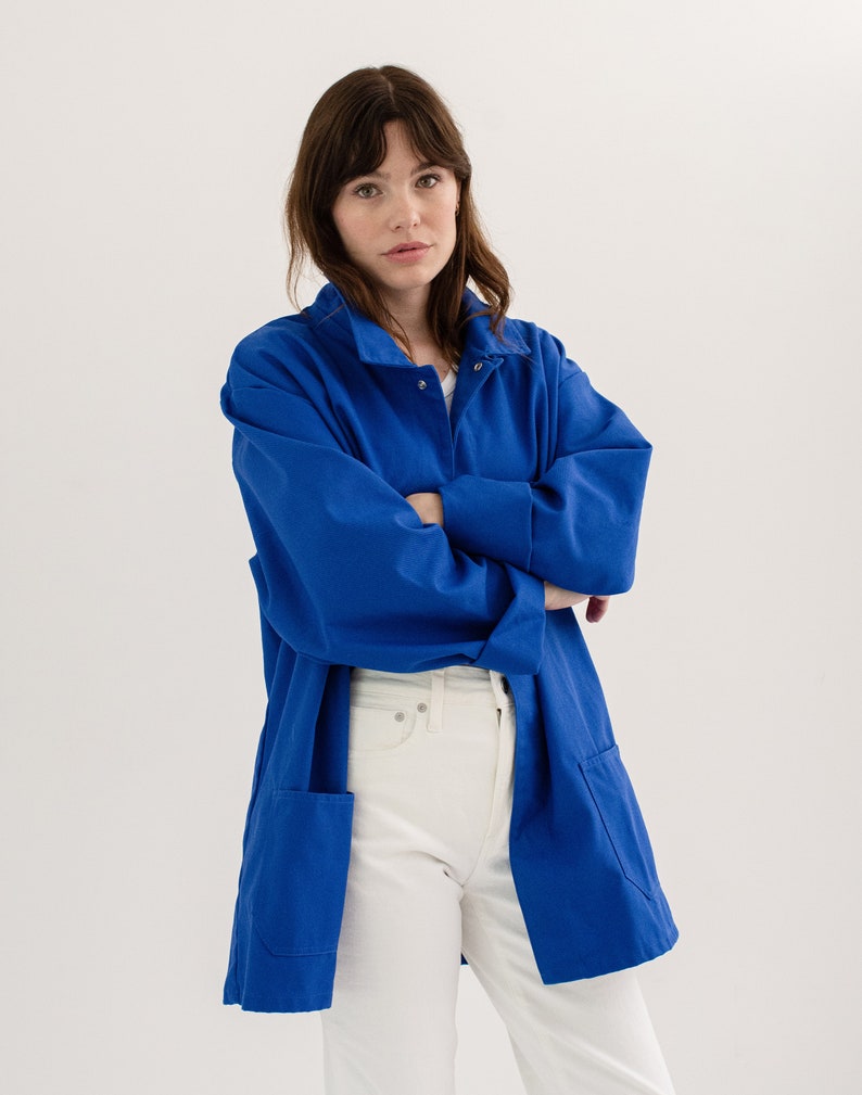 May include: A woman wearing a long, royal blue work jacket with a white button-down shirt underneath. She is wearing white jeans and has her arms crossed.
