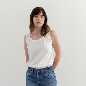 May include: A woman wearing a white tank top and blue jeans. She is standing in front of a white background.