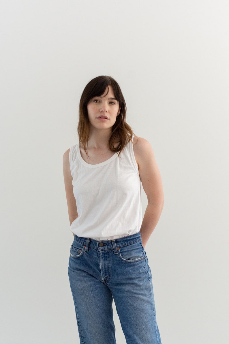 May include: A woman wearing a white tank top and blue jeans. She is standing in front of a white background.