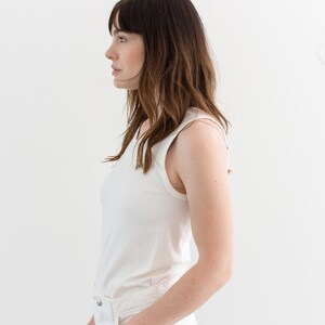 May include: A woman wearing a white sleeveless top and white jeans. She is standing in front of a white wall.