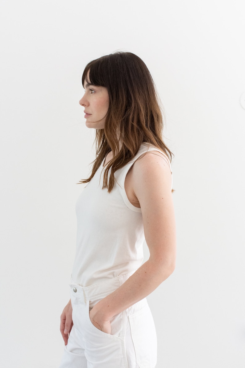May include: A woman wearing a white sleeveless top and white jeans. She is standing in front of a white wall.