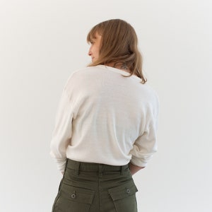 May include: A long-sleeved, off-white crew neck shirt paired with olive green trousers. The trousers feature a high-waisted design with two prominent rectangular back pockets, each secured with a single dark button at the center of the flap.