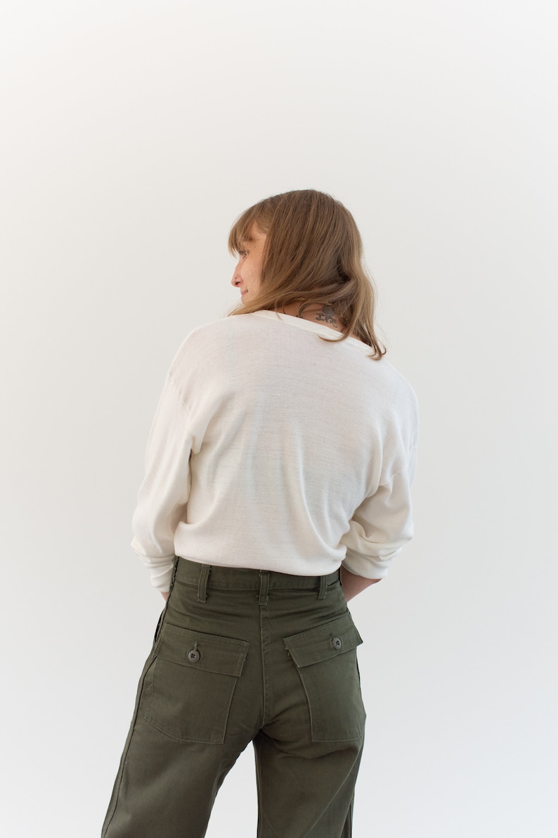 May include: A long-sleeved, off-white crew neck shirt paired with olive green trousers. The trousers feature a high-waisted design with two prominent rectangular back pockets, each secured with a single dark button at the center of the flap.