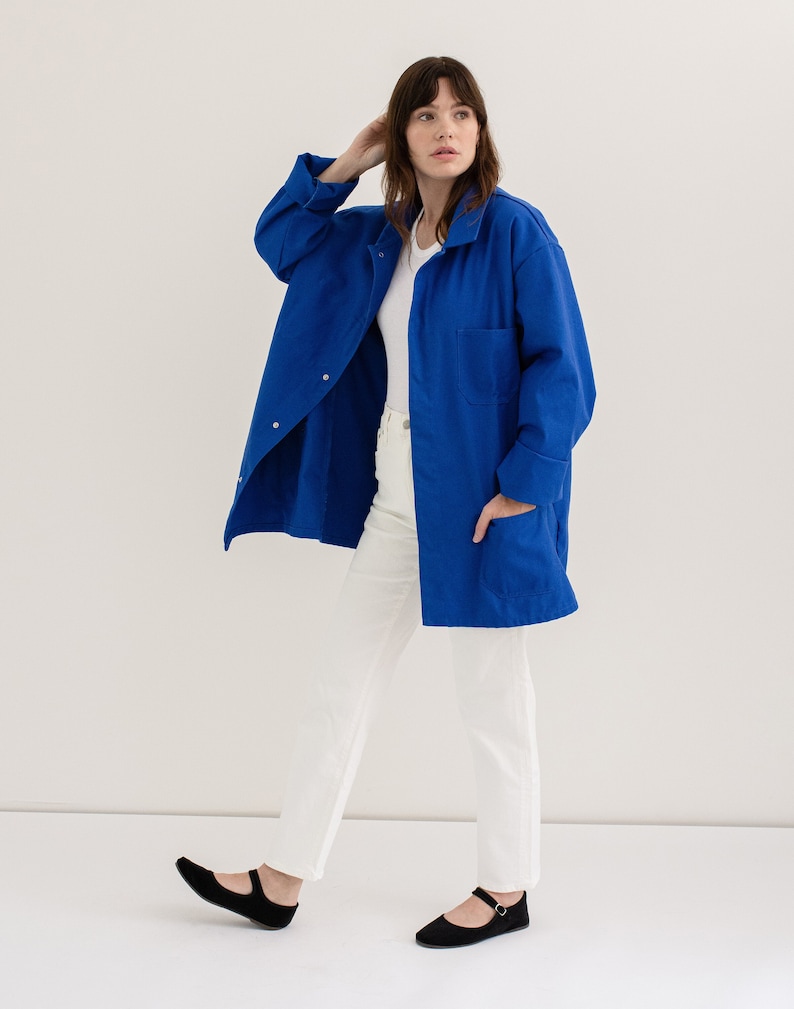 May include: A woman wearing a long, royal blue utility jacket with a white t-shirt and white pants. She is wearing black flats with a strap.