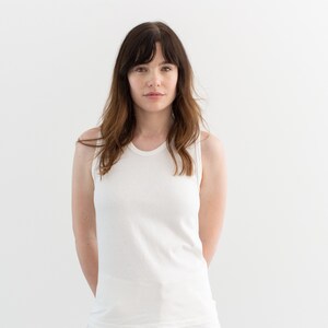May include: A woman wearing a white tank top and white pants. She is standing in front of a white background.