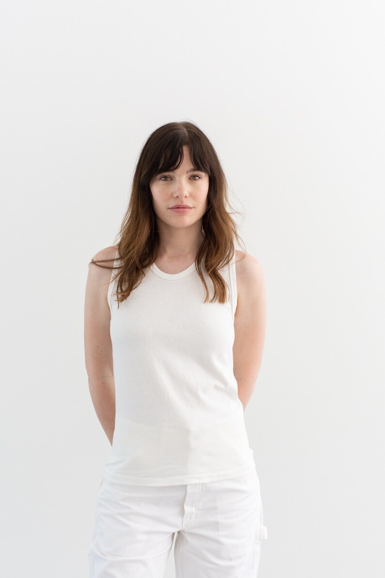 May include: A woman wearing a white tank top and white pants. She is standing in front of a white background.