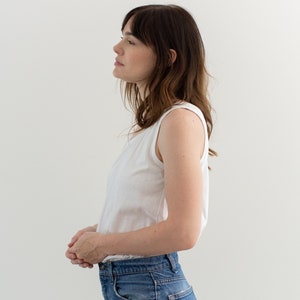 May include: A woman wearing a white tank top and blue jeans. She is standing in front of a white wall.