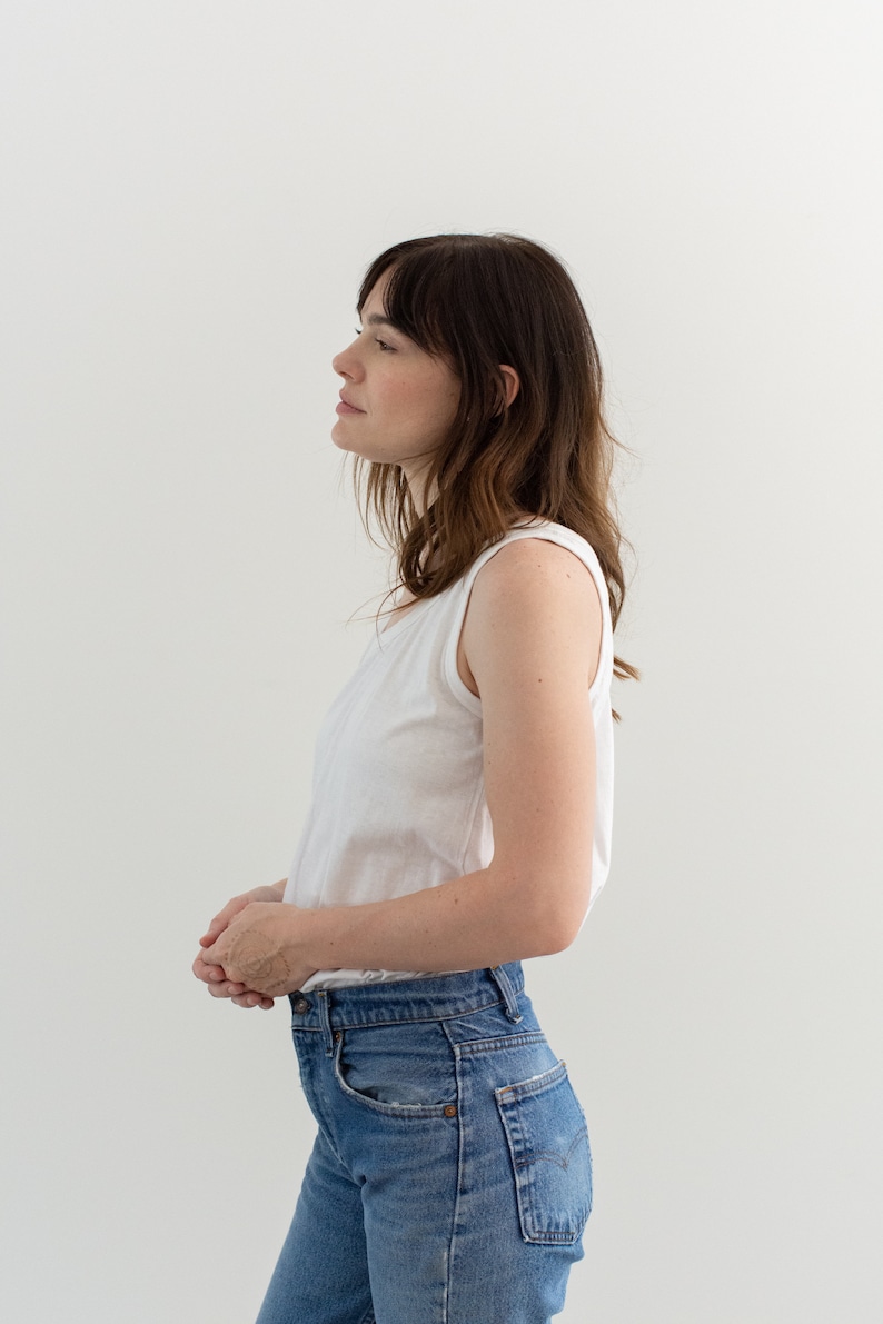May include: A woman wearing a white tank top and blue jeans. She is standing in front of a white wall.