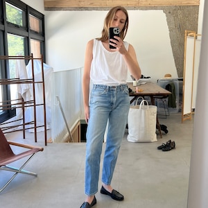 Puede incluir: Una persona lleva un top blanco sin mangas y unos vaqueros azules claros con mocasines negros. La persona est&aacute; de pie en una habitaci&oacute;n con suelo de madera y una gran ventana en el fondo.