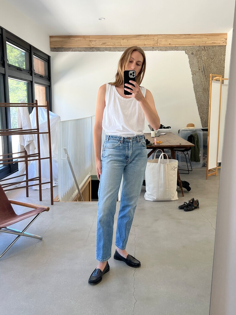 Puede incluir: Una persona lleva un top blanco sin mangas y unos vaqueros azules claros con mocasines negros. La persona est&aacute; de pie en una habitaci&oacute;n con suelo de madera y una gran ventana en el fondo.