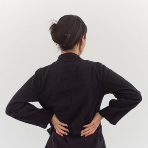 Op de afbeelding: Een vrouw draagt een zwart blazer met lange mouwen. Ze staat met haar handen op haar heupen en haar rug naar de camera.