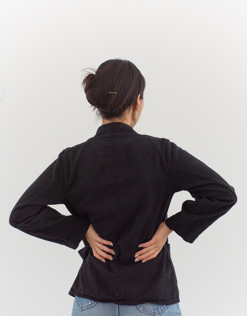 Op de afbeelding: Een vrouw draagt een zwart blazer met lange mouwen. Ze staat met haar handen op haar heupen en haar rug naar de camera.