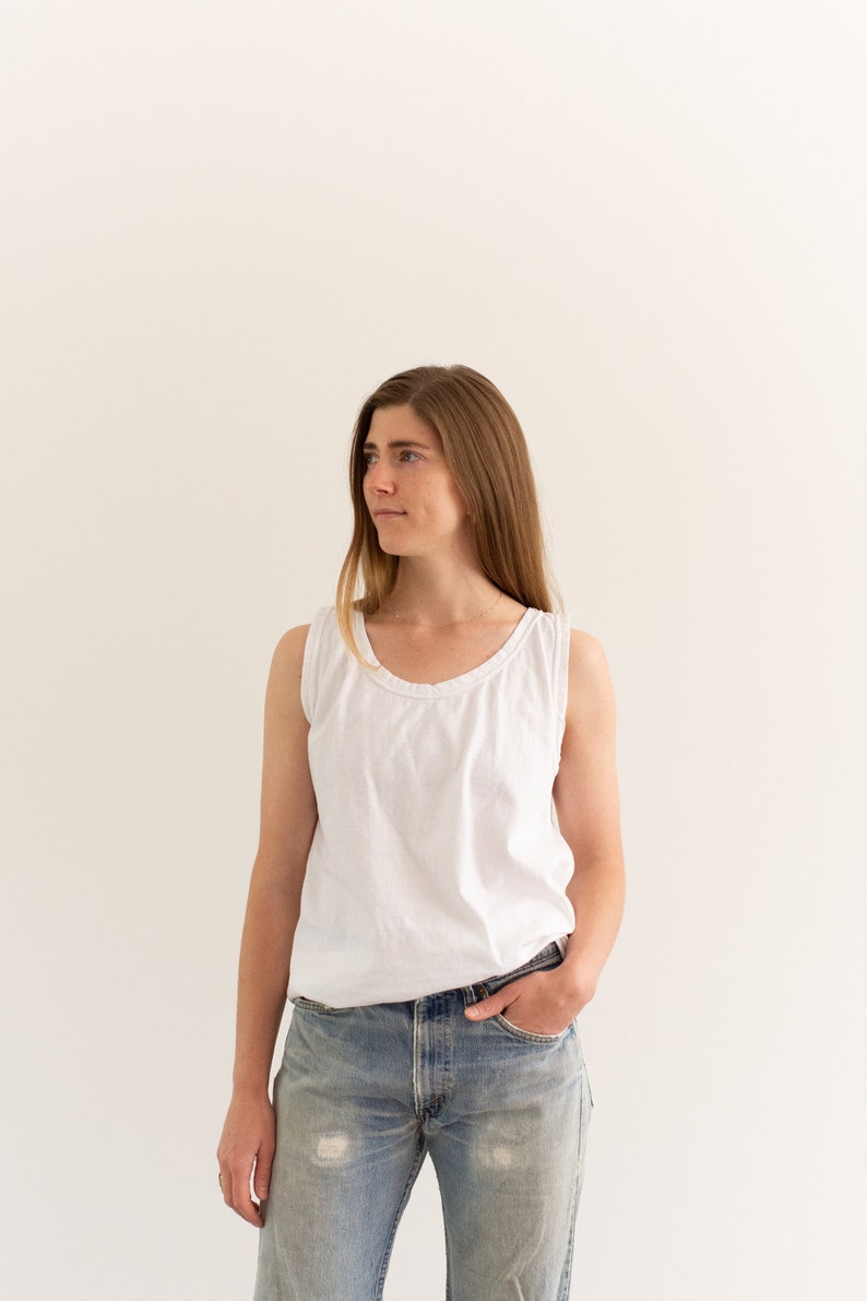 May include: A woman wearing a white tank top and light blue jeans. She is standing in front of a white wall.