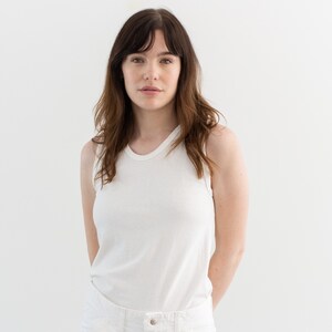 May include: A woman wearing a white tank top and white jeans. She is standing in front of a white background.