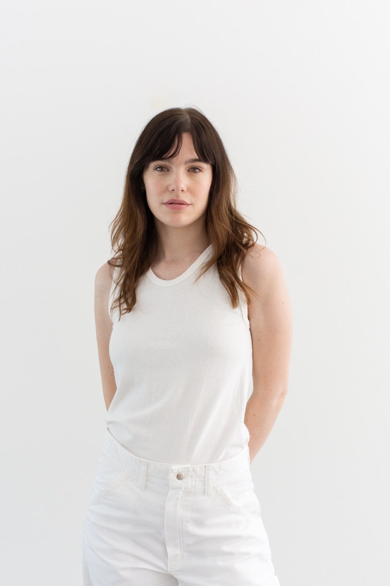 May include: A woman wearing a white tank top and white jeans. She is standing in front of a white background.