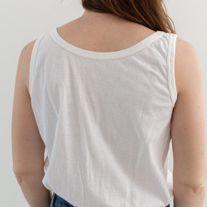 May include: A white sleeveless tank top with a scoop neck. The tank top is worn with blue denim jeans.