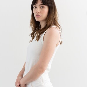 May include: A woman wearing a white tank top and white pants stands against a white background.