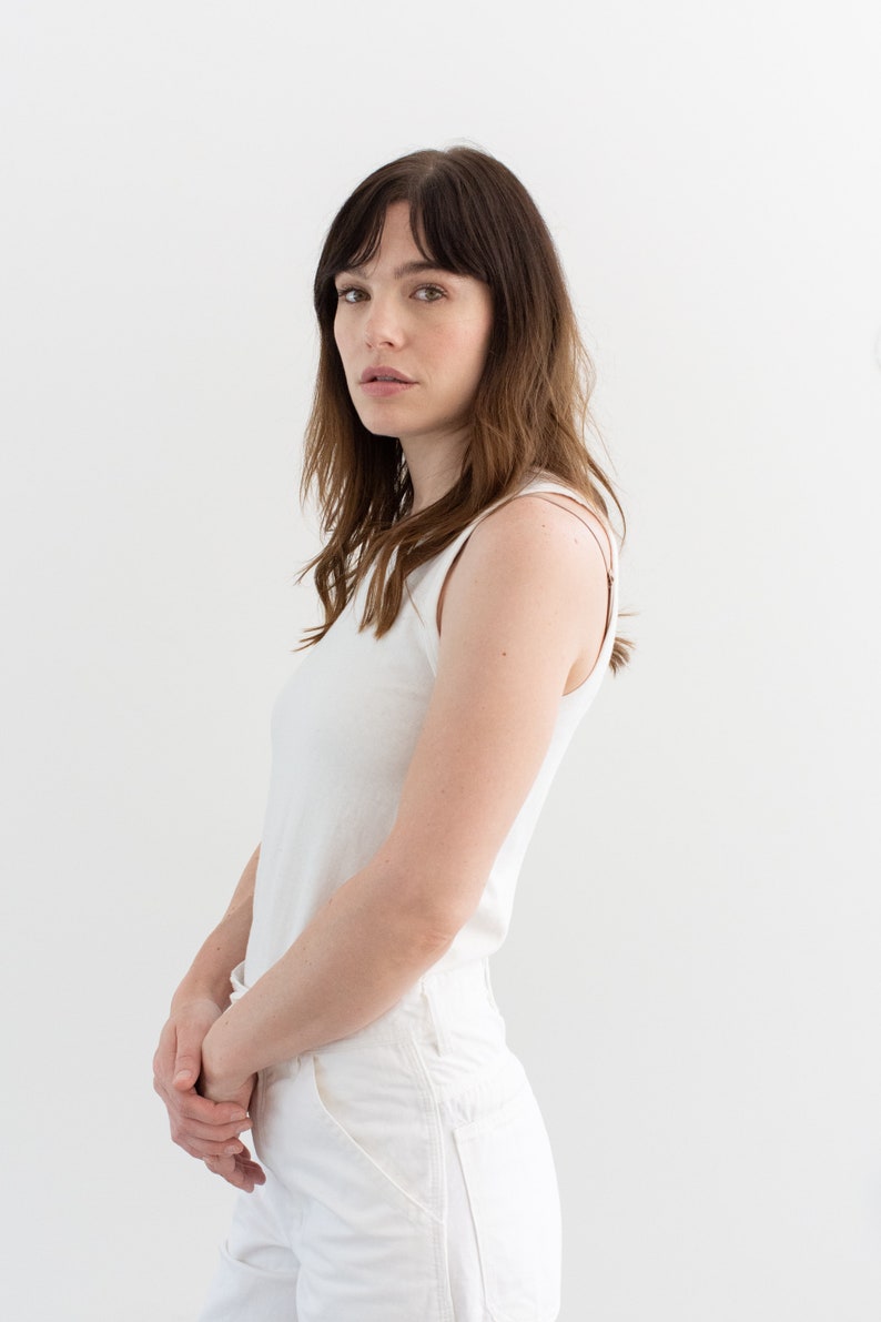 May include: A woman wearing a white tank top and white pants stands against a white background.