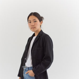 Op de afbeelding: Een vrouw draagt een zwart blazer over een wit t-shirt en een blauwe jeans. Ze staat voor een witte achtergrond.
